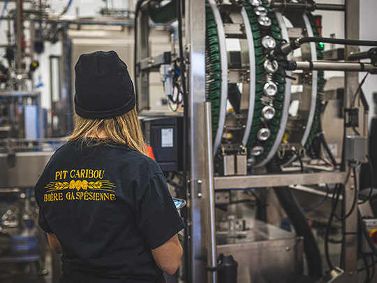 Woman seen from behind wearing a black t-shirt from the Pit Caribou brewery and facing the automated canning line.