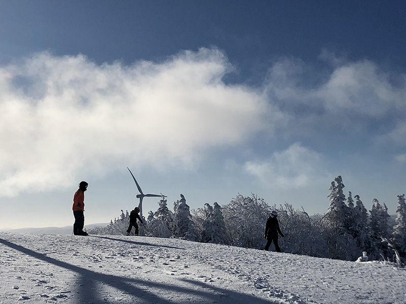 A family is all geared up for snowboarding on a snowy trail at Mount Miller; a wind turbine is visible in the background