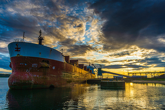 A transoceanic vessel docked in an industrial port under a dramatic sky.