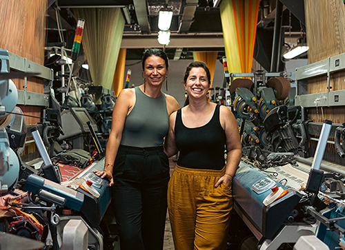 Mélanie Bernard and Trisha Pitura pose in the middle of the MINI TIPI production workshop, with industrial weaving machines on either side of the photo.